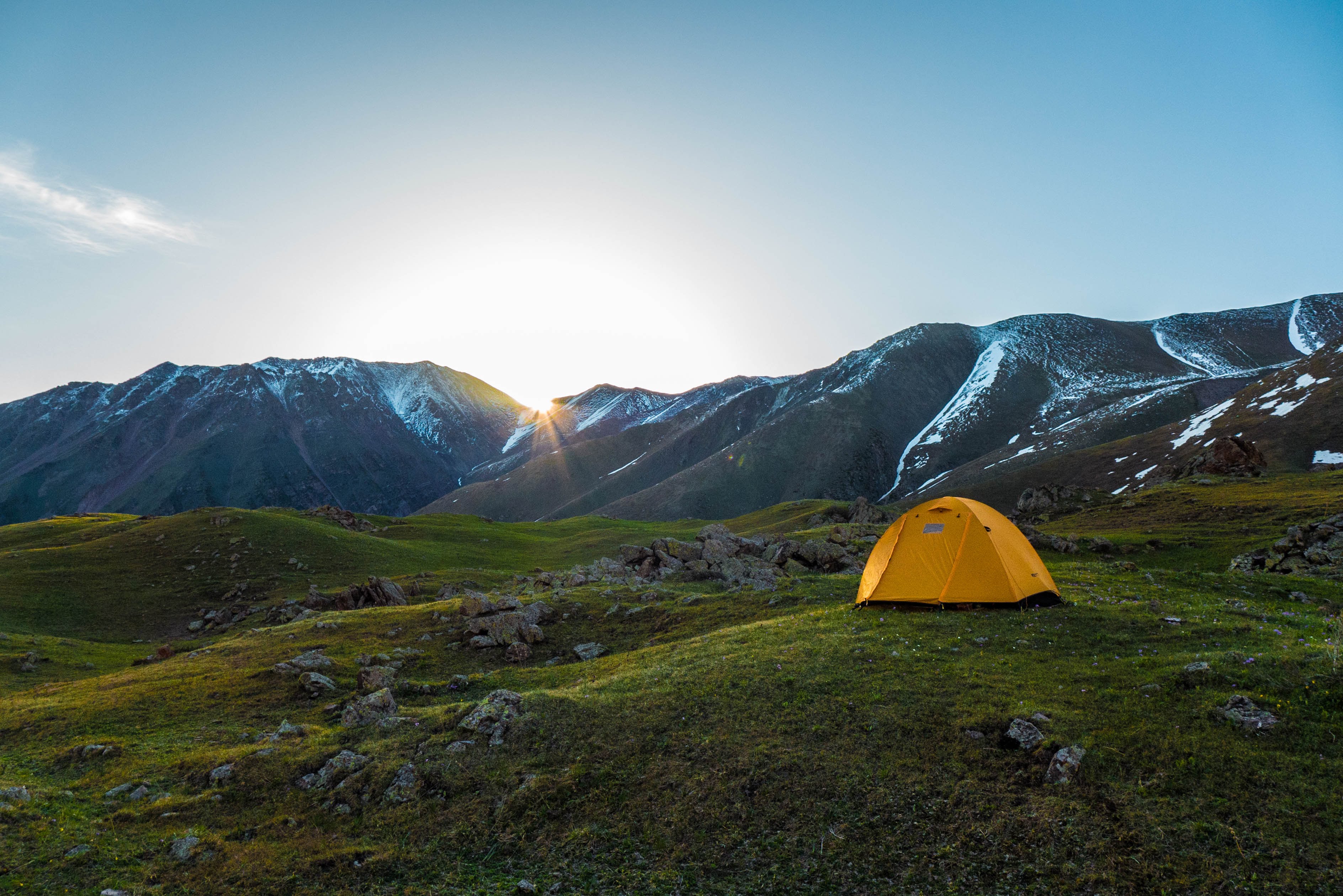 A bright yellow tent pitched on grassy terrain in a high alpine valley with snow-capped mountains in the background at sunrise.